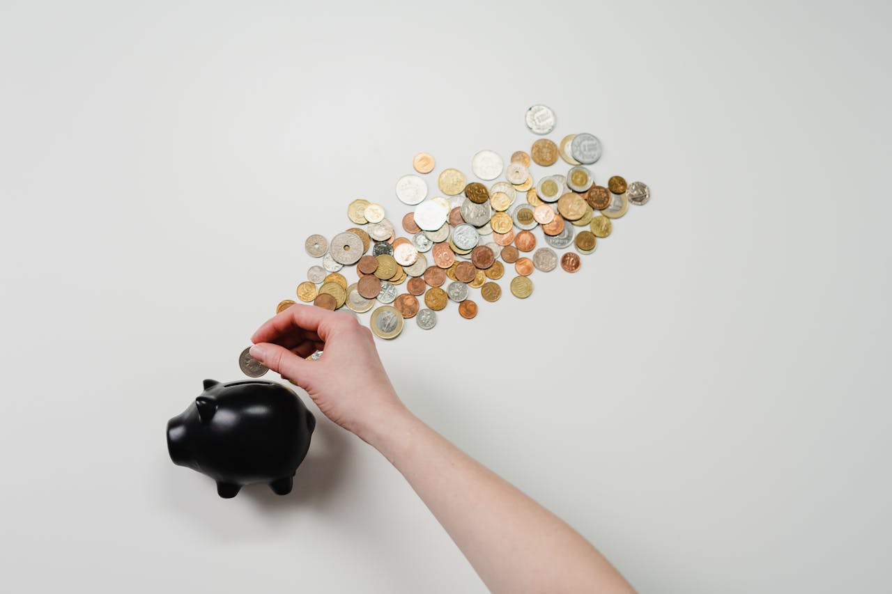 heros-img Person placing coin into a black piggy bank with scattered coins, symbolizing savings and finance.
